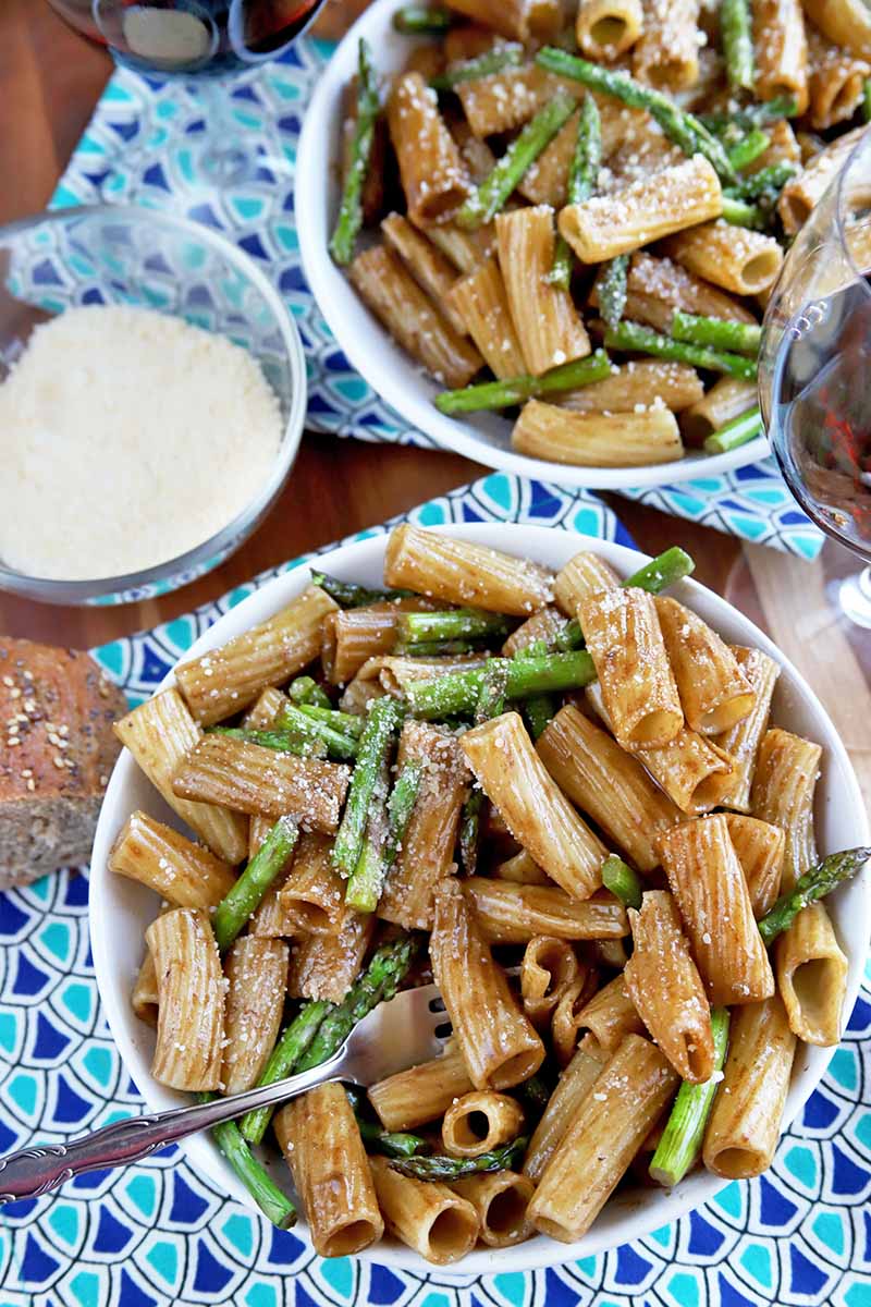 Vertical overhead shot of two white bowls of pasta with asparagus and a brown balsamic vinegar sauce, with a small glass bowl of grated cheese and a fork in the bowl in the foreground, with two glasses of wine mostly cropped out of the top and right sides of the frame, on a dark and light blue cloth on a brown wood surface.
