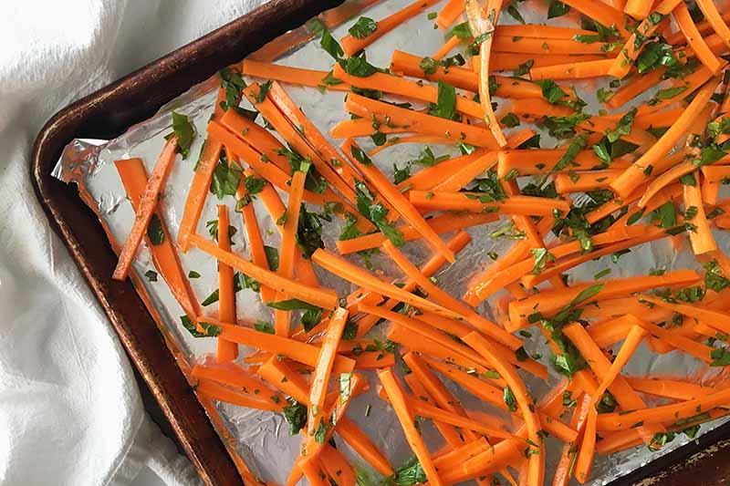 Horizontal image of raw carrot sticks mixed with herbs on a sheet pan.