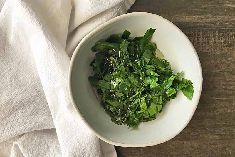 Horizontal image of chopped fresh herbs in a white bowl next to a white towel.