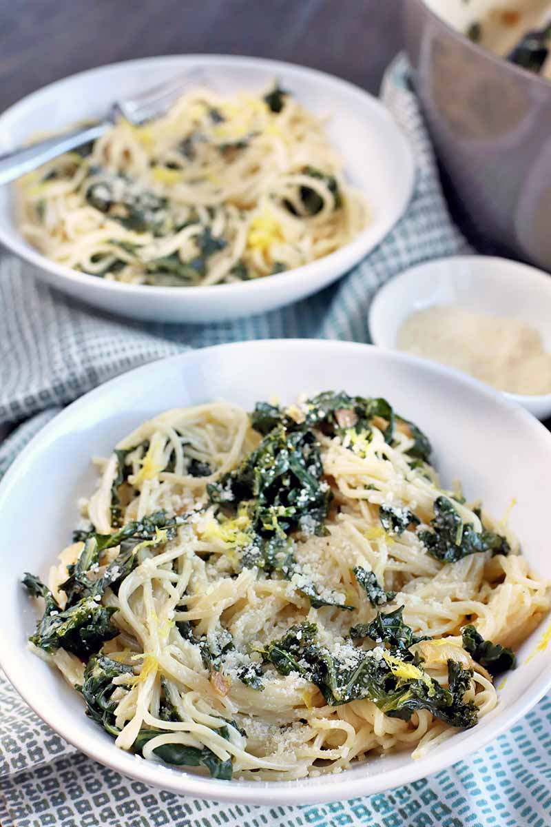 Two shallow white bowls of cooked pasta and green kale topped with cheese, with a small white bowl of grated Parmesan and a stainless steel serving bowl, on a dark brown wood surface with a white and light blue cloth.