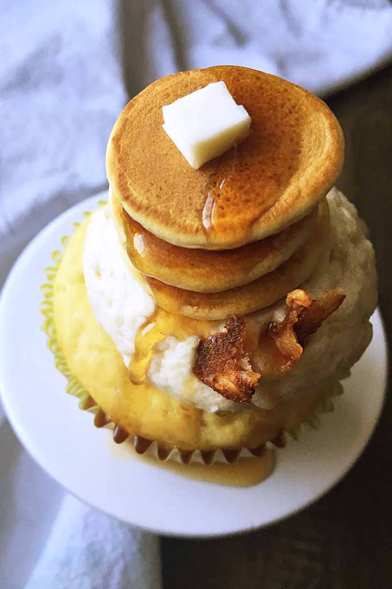 Vertical image of a cupcake topped with a stack of pancakes, a pat of butter, bacon, and syrup.