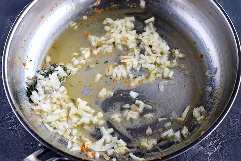 Onions are being sauteed in butter in a large metal frying pan, on a gray slate surface.