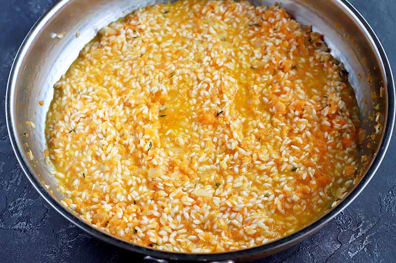 Rice cooks in an orange liquid in a large metal frying pan, with onions and thyme and a few flecks of carrot, on a dark gray slate surface.