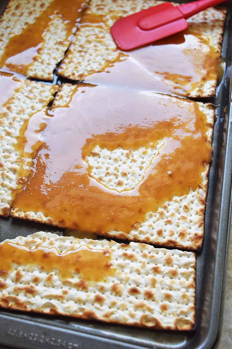Oblique vertical shot of a red rubber spatula being used to spread caramel sauce on pieces of matzo in a rimmed metal baking pan.