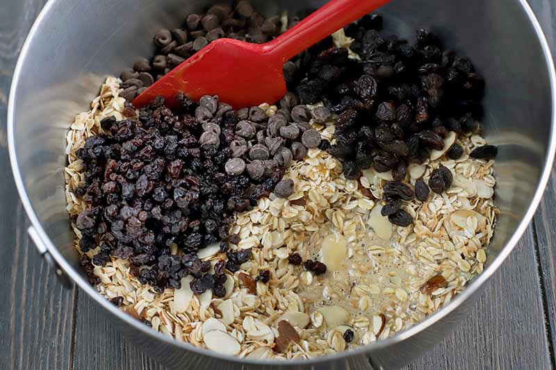 A red rubber spatula stirs a mixture of dried fruit, chocolate chips, almonds, shredded toasted coconut, and oats in a stainless steel bowl, on a dark brown wood surface.