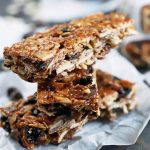 Three stacked homemade granola bars stacked in the foreground with more in soft focus in the background, on pieces of crumpled white parchment paper, on a dark brown wood surface with scattered oats and chocolate chips.