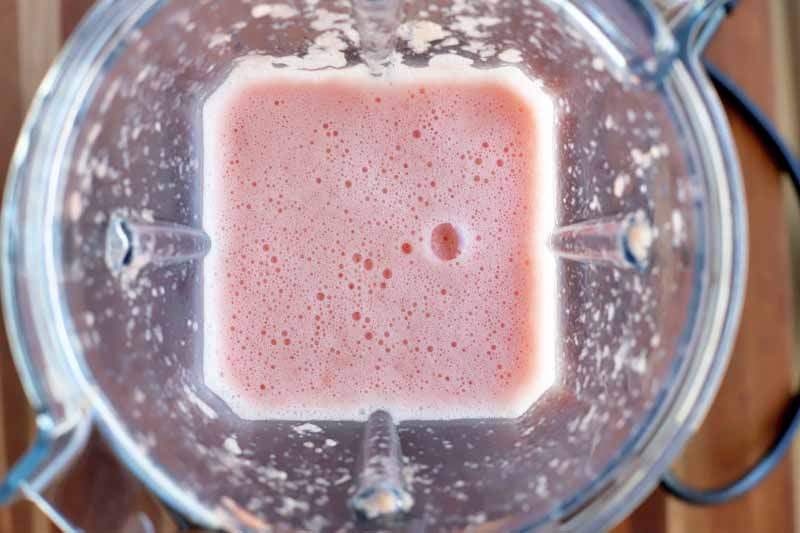 Vertical overhead image of a clear plastic countertop blender pitcher with a frothy pink blended fruit mixture at the bottom, with a black power cord to the right, on a brown wood surface.