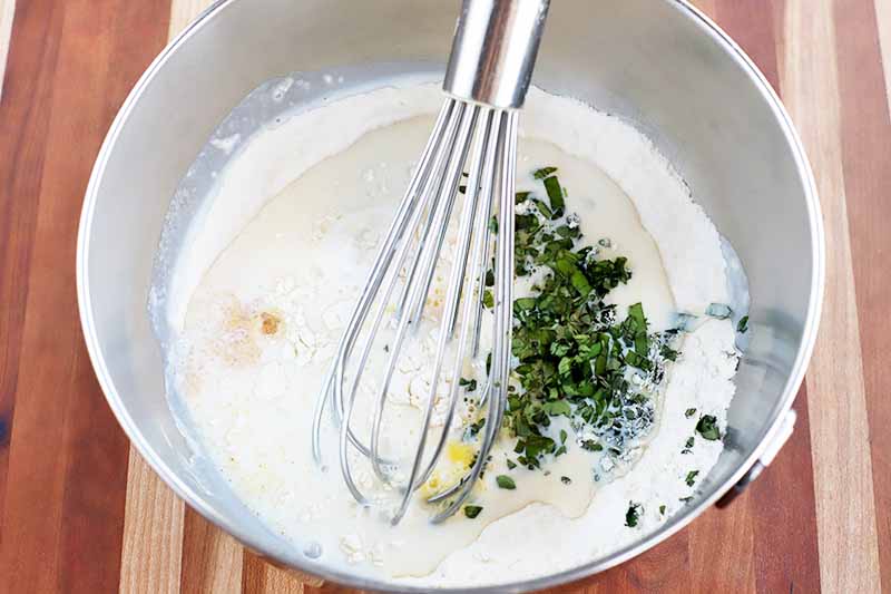 A wire whisk whips chopped fresh herbs into a batter mixture in a stainless steel bowl, on a beige and brown wood table.