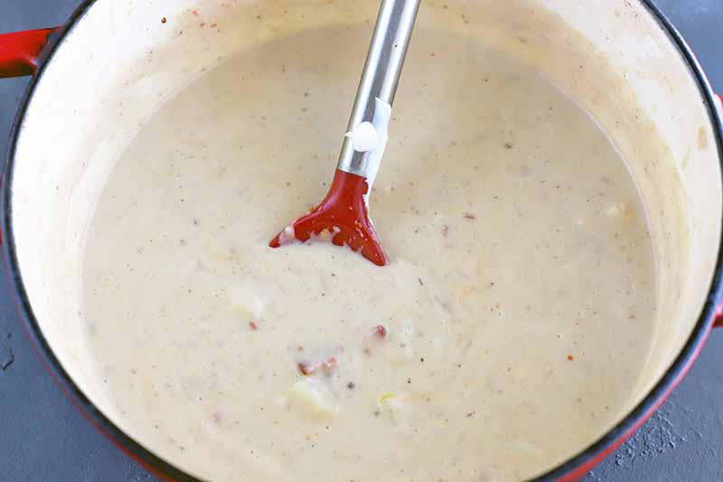 A red silicone slotted spoon with a silver metal handle is being used to stir potato soup in a large enameled cast iron Dutch oven with red handles, on a gray slate surface.
