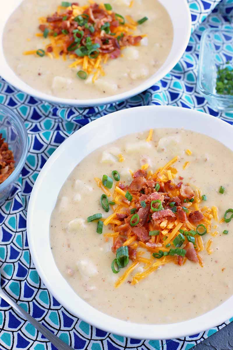 Vertical closeup closely cropped image of two white bowls of loaded baked potato soup with a variety of garnishes, with two small glass bowls of crumbled bacon and chopped chives, on a light and dark blue patterned cloth surface.