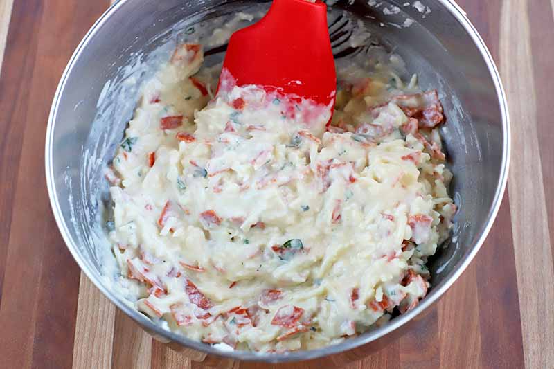 A red silicone spatula stirs chopped pepperoni into a batter mixture with fresh herbs and cheese in a stainless steel mixing bowl, on a striped wood table.