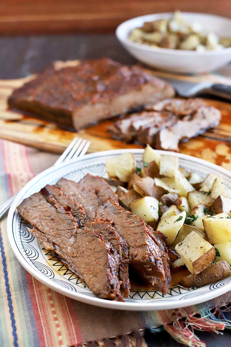 Vertical oblique shot of a plate of sliced brisket and roasted potatoes, with more of each on a wooden serving platter and in a white bowl in the background, on a striped multicolored cloth with fringe, on a table, with a brown background.
