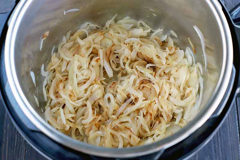 Horizontal image of thinly sliced onions browning in the bottom of a stainless steel Instant Pot insert, on a dark brown wood surface.