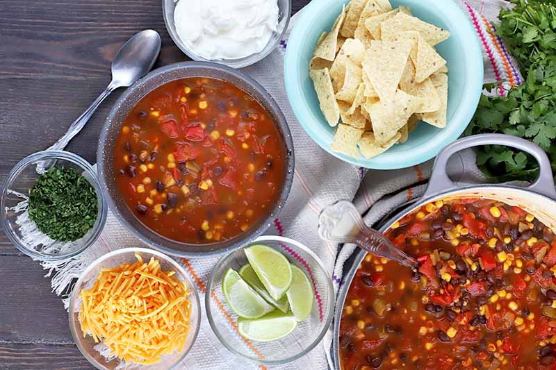 Overhead shot of a pot and a bowl of bean, corn, and tomato soup with a blue bowl of tortilla chips, four glass bowls of sour cream, chopped herbs, shredded cheese, and lime wedges, on a white cloth with fringe, a spoon to the left, and a bunch of fresh cilantro to the right, on a dark brown wood surface.