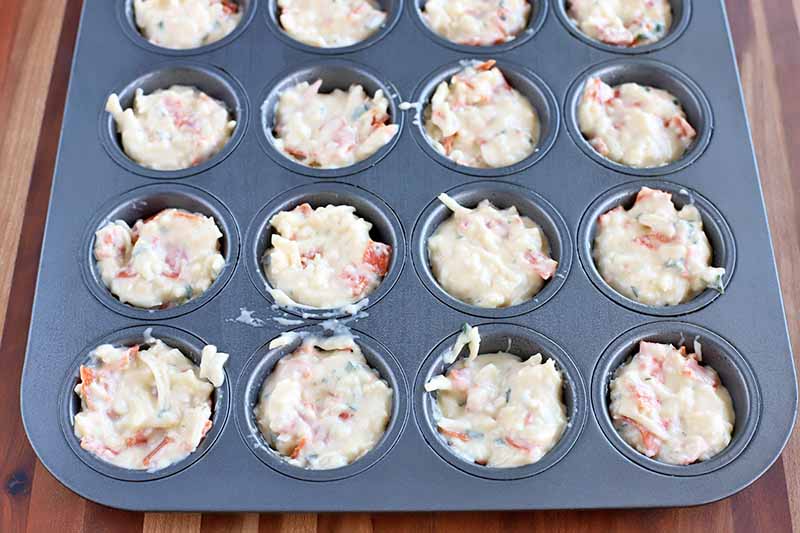 Batter has been portioned into the wells of a metal mini muffin tin, on a brown background.