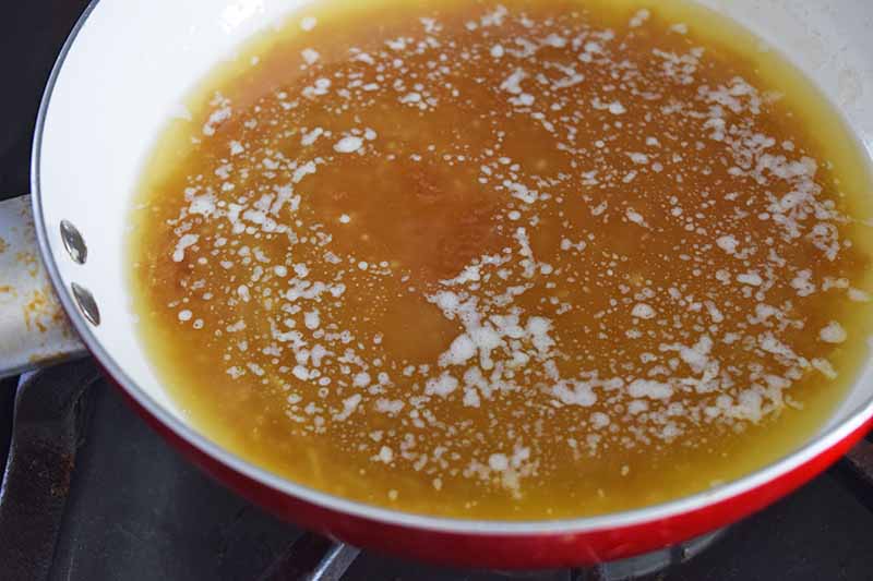 A red and white frying pan of homemade caramel sauce being made with brown sugar and frothy melted butter, on a gas stove.