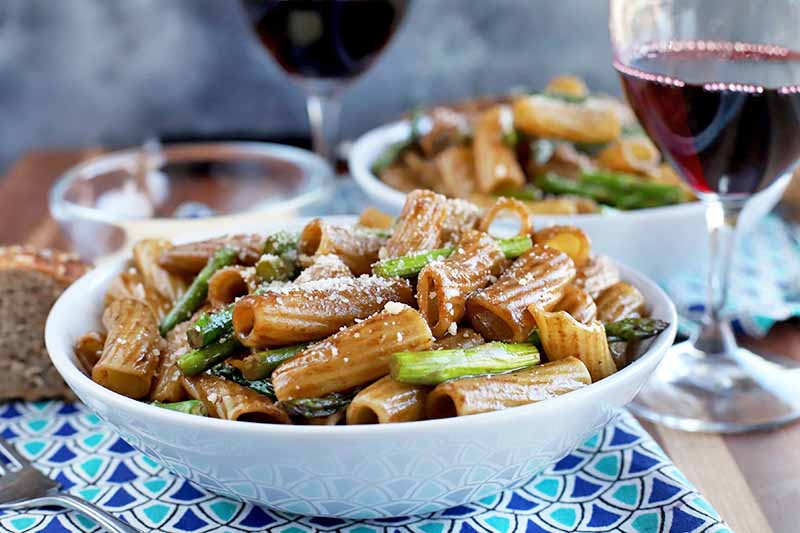 Horizontal head-on image of two white bowls of pasta with asparagus and a brown sauce, with a small glass bowl of grated Parmesan and two glasses of red wine, on a dark and light blue cloth with a triangular pattern on a brown table, with a mottled gray background.