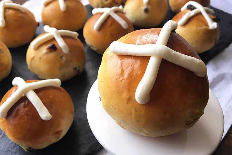 Horizontal closeup image of a homemade hot cross bun on a white stand, surrounded by more in the background.