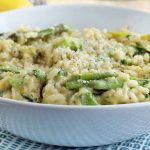 Horizontal image of a large white bowlful of asparagus risotto, on a white and blue checkered cloth on a brown wood surface with a sliced lemon in the background.