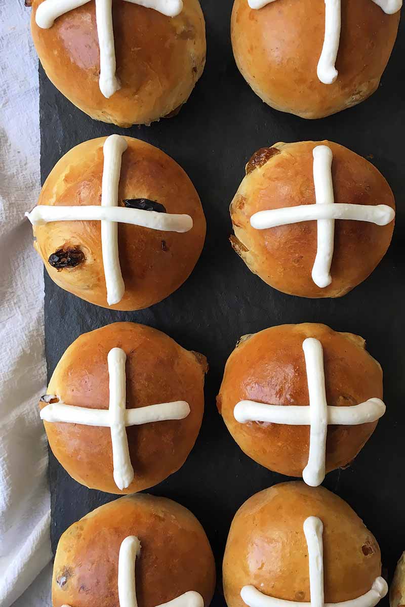 Vertical top-down image of two columns of hot cross buns on a dark slate surface next to a white towel.