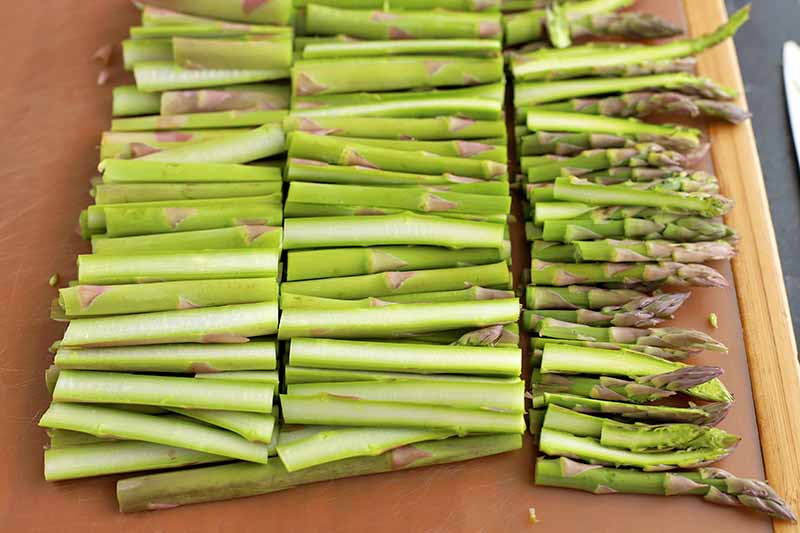 A brown cutting board is topped with rows of asparagus that have been halved lengthwise and then cut width-wise into thirds.