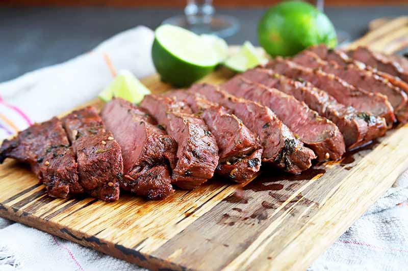 Horizontal image of sliced meat on a wooden cutting board with a whole lime and sliced limes.
