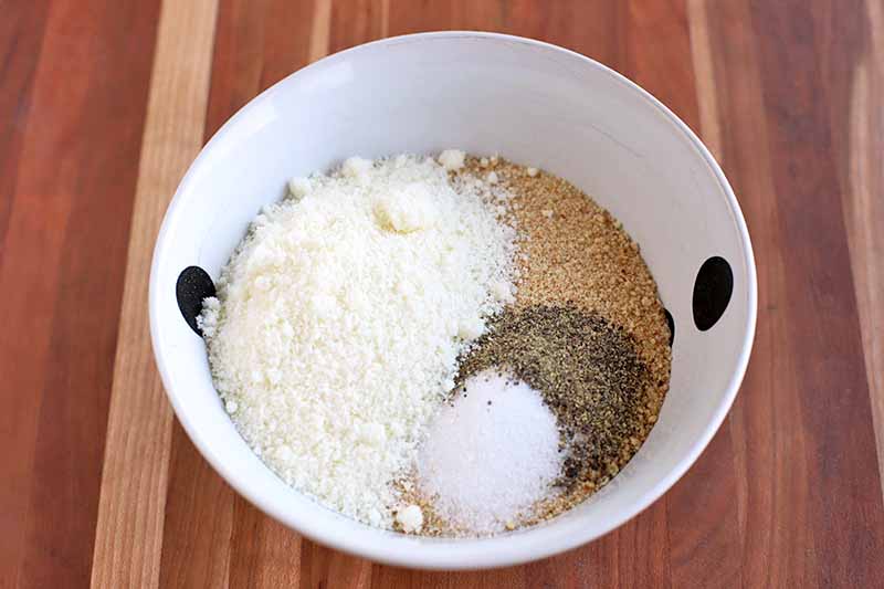 Oblique overhead horizontal shot of a white bowl with two black spots, with breadcrumbs at the bottom topped with small piles of grated cheese, salt, and ground black pepper, on a brown wood surface.
