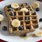 Two buckwheat buttermilk waffles on a plate with sliced banana and fresh blueberries on top, on a red cloth background.