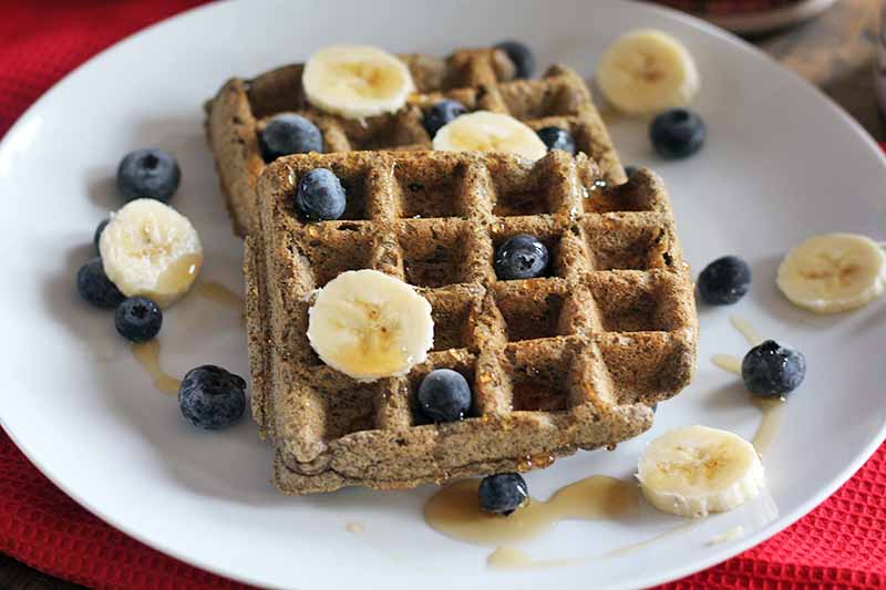 Two buckwheat buttermilk waffles on a plate with sliced banana and fresh blueberries on top, on a red cloth background.