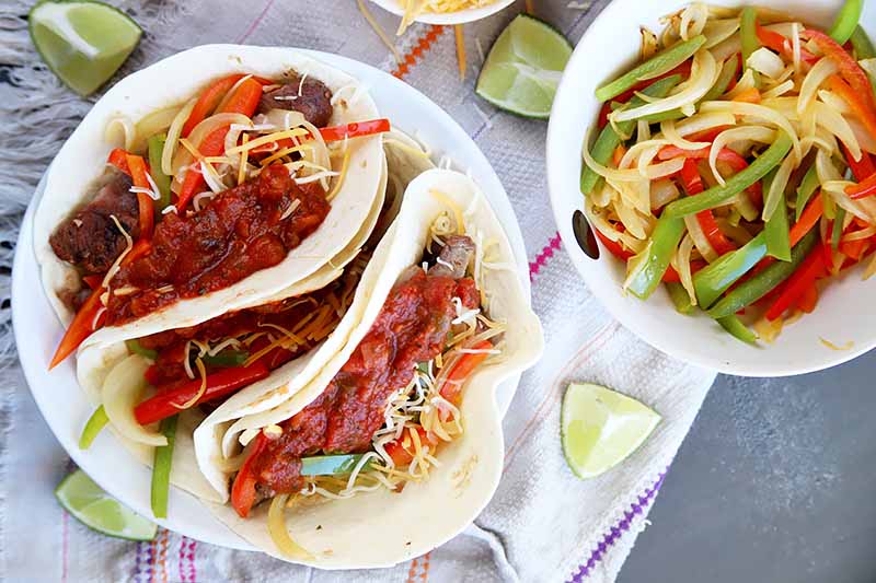 Horizontal top-down image of tacos on a white plate, next to cut limes and a plate of grilled vegetables.