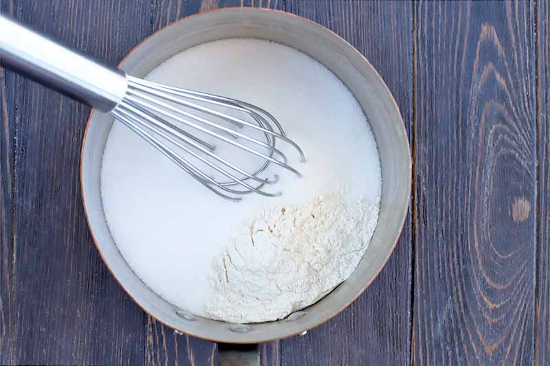A wire whisk stirs flour into a mixture of milk and sugar in a metal saucepan, on a dark brown wood surface.