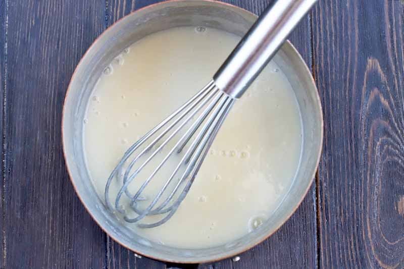 A wire whisk stirs a milky mixture in a metal saucepan, on a brown wood table.
