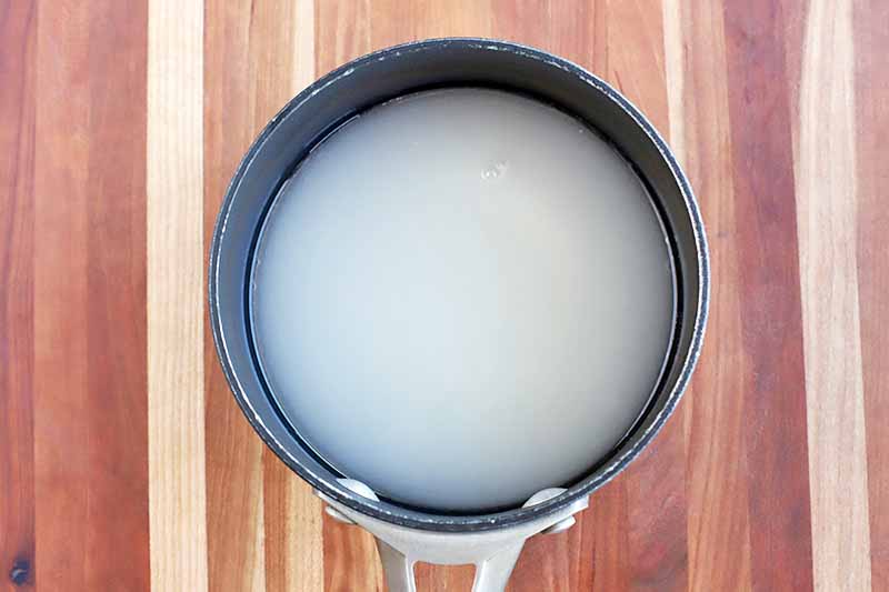 Overhead shot of a sugar syrup mixture in a saucepan, on a brown and beige wood surface with vertical stripes.