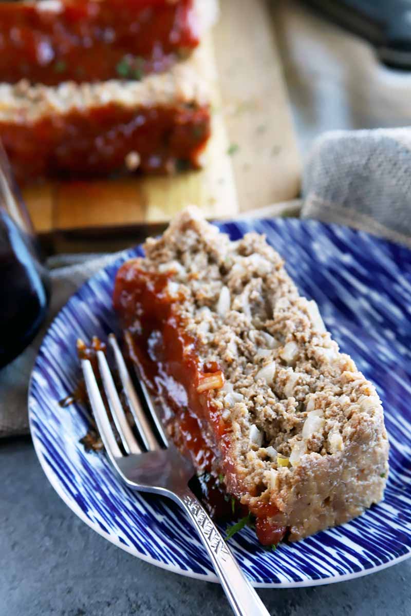Vertical image of one slice of meatloaf on a blue plate with a metal fork.