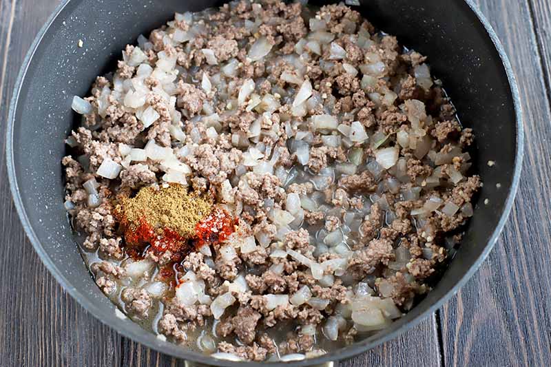 Overhead closely cropped shot of a large nonstick frying pan of cooked ground beef and chopped onions, with a small pile of ground cumin, chili, and other spices on top, on a dark brown wood surface.