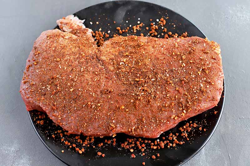 Overhead horizontal shot of a cut of raw beef on a black ceramic plate, coated with a spice rub mixture, on a gray stone surface.