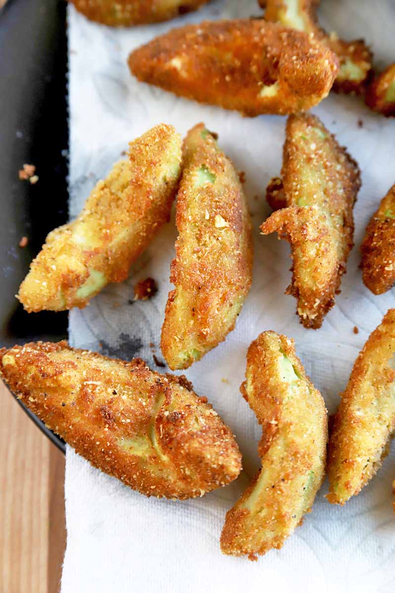 Vertical overhead shot of golden, crispy, batter-dipped fried avocado wedges, on a white cutting board on top of a wood surface.