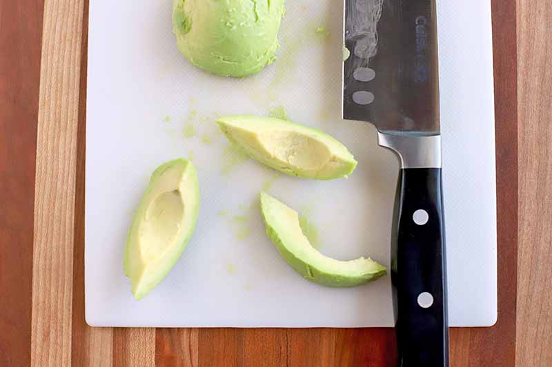 Horizontal closely cropped overhead shot of a white cutting board topped with a peeled avocado, with one half at the top of the frame and three wedge slices at the bottom, next to a knife with a black handle, on a beige and brown wood surface with vertical stripes.