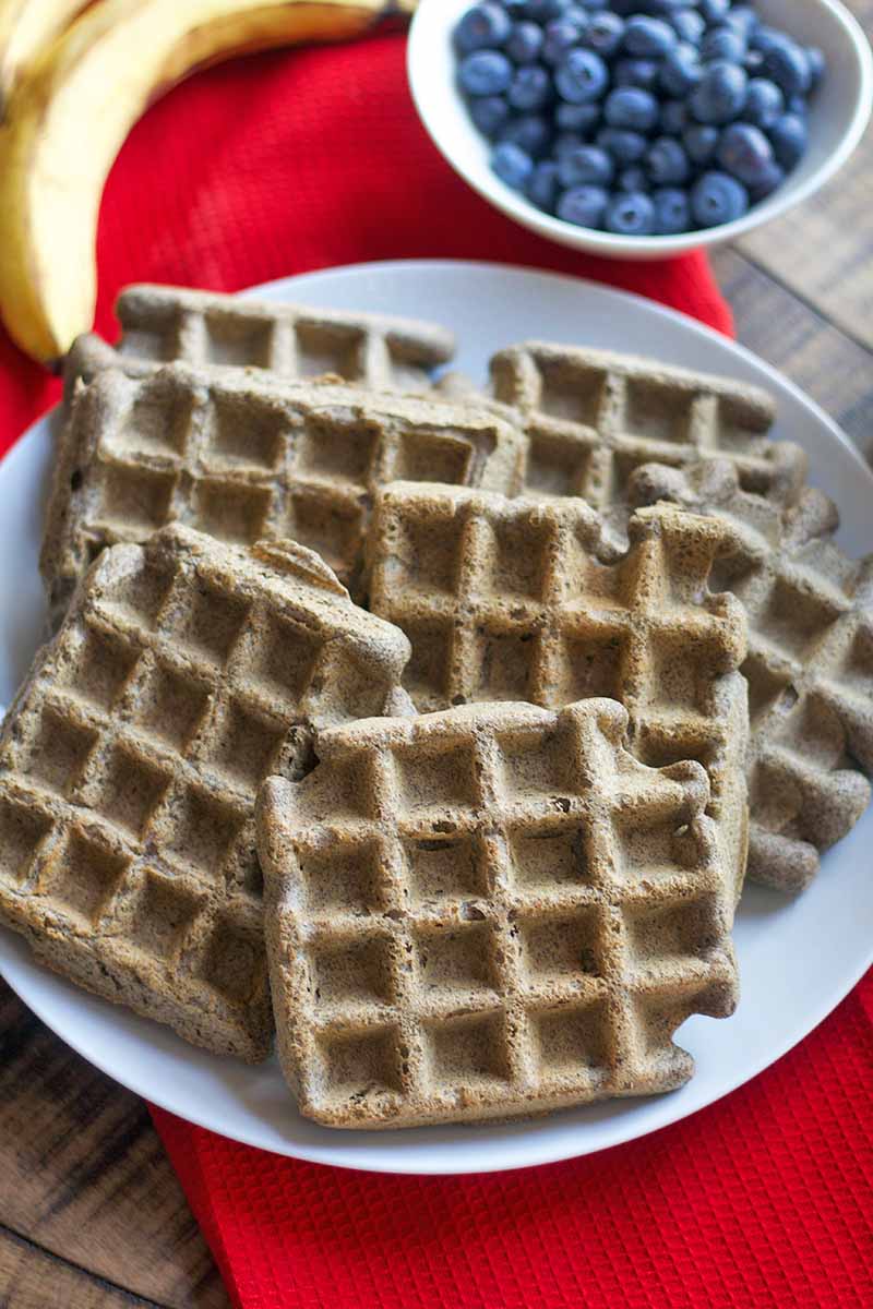 Six freshly made buckwheat waffles are arranged on a white plate, with a small white bowl of blueberries and two yellow bananas, on a red cloth on top of a brown wood surface, shot from above.