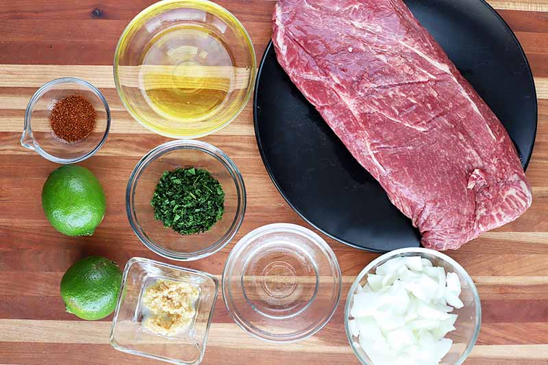 Horizontal image of a whole steak on a black plate next to assorted prepped ingredients.