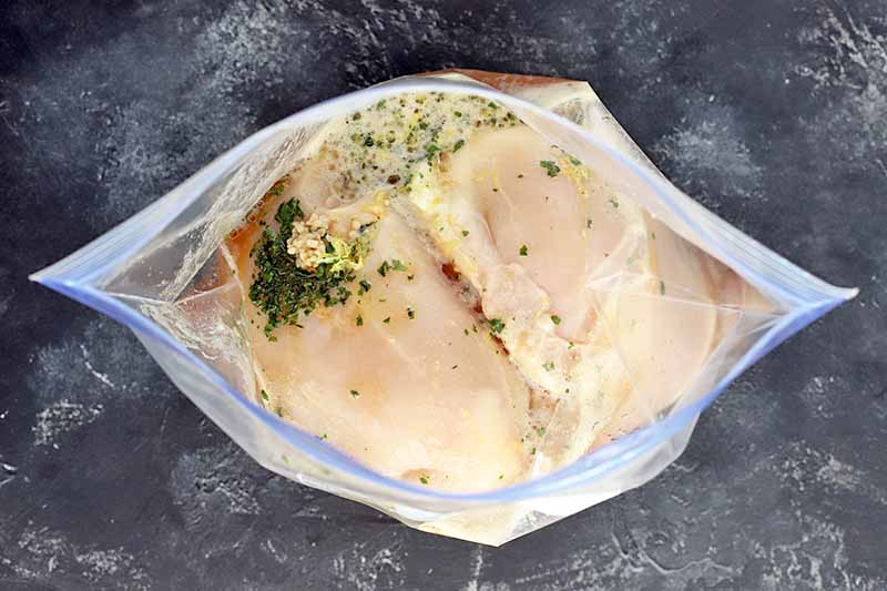 Overhead horizontal shot of a large zip-top bag filled with raw chicken breasts and a marinade mixture of garlic, fresh herbs, citrus juice, and lager beer, on a gray and white surface.