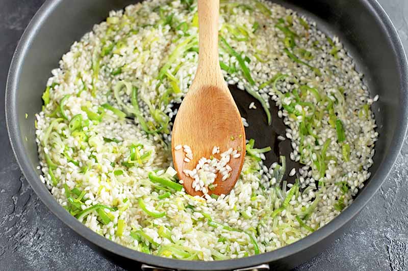 Horizontal image of a large frying pan of arborio rice, leeks, and garlic, being stirred with a wooden spoon, on a gray slate background.