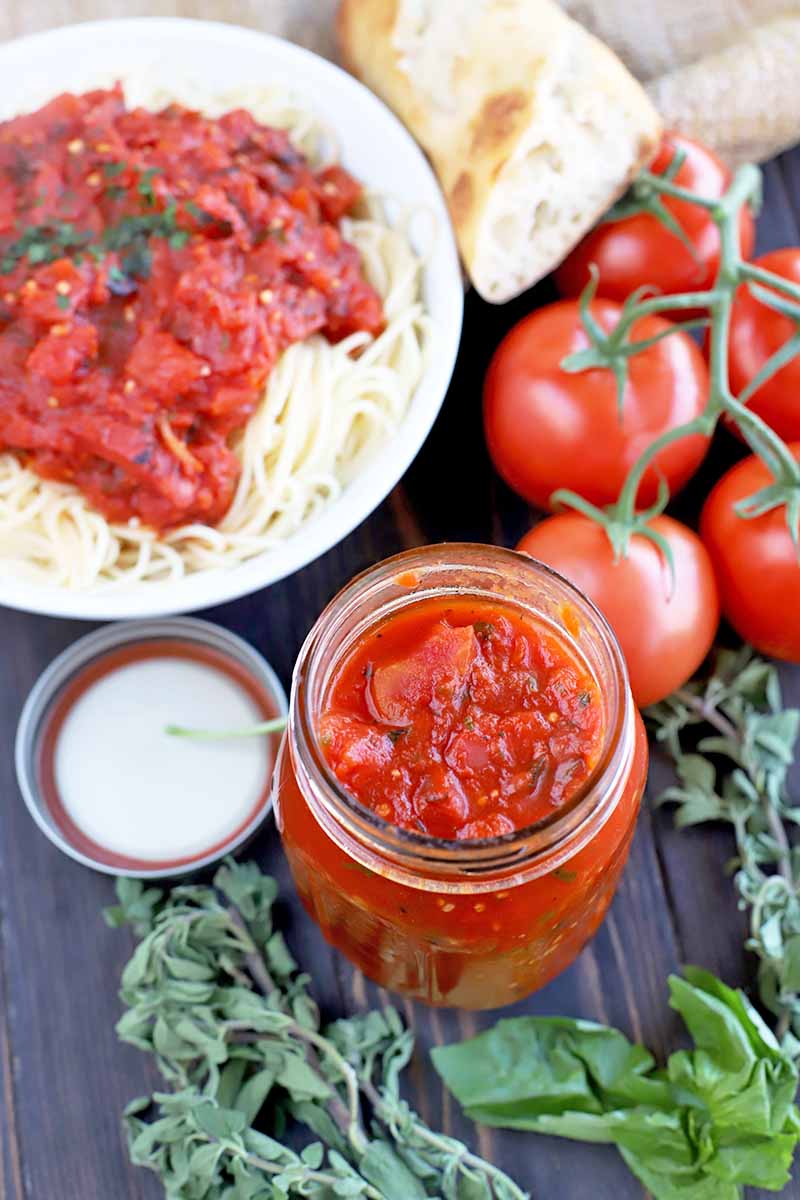 Vertical overhead image of a glass mason jar of marinara with tomatoes on the vine, a white plate of pasta and sauce, sprigs of fresh oregano and basil, and a hunk of crusty bread, on a folded piece of burlap on a dark brown wood surface.