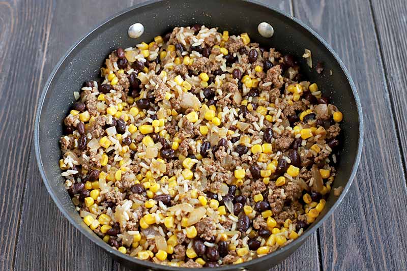 Overhead shot of a large fying pan of ground beef, beans, corn, and other ingredients, on a dark brown wood table.