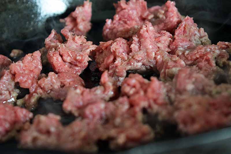 Raw, pink ground beef is cooking in a cast iron pan. Horizontal image with selective focus.