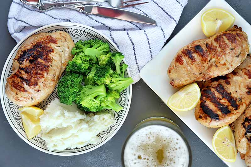 Overhead shot of a round white plate and a rectangular serving platter of grilled chicken with lemon, with mashed potatoes and broccoli on the side of the dinner plate, a white cloth napkin with blue stripes topped with silverware, and a mug of foamy beer, on a gray slate background.