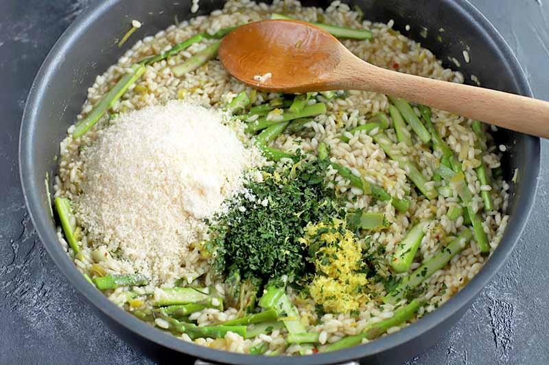 Horizontal image of a large frying pan of rice, leeks, and asparagus, with small piles of grated cheese, parsley, and lemon zest on top, with a wooden spoon, on a gray slate background.