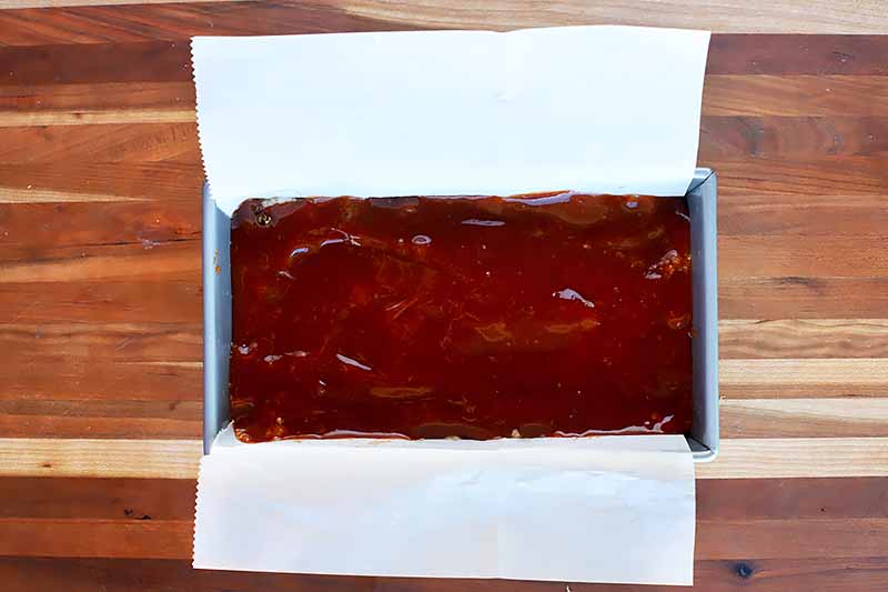 Horizontal image of a metal loaf pan with a food item topped with a ketchup glaze.