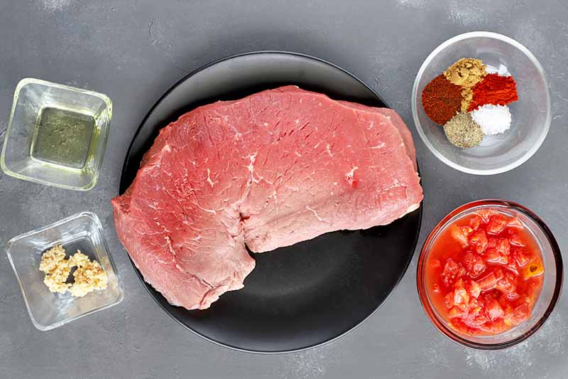 Overhead horizontal shot of a cut of beef sirloin on a black ceramic plate, with two square glass dishes of salt and pepper and minced garlic to the left, and two round glasses dishes of various spices and canned diced tomatoes to the right, on a gray surface.
