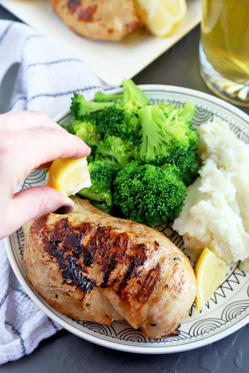 A hand with manicured dark purple nails squeezes a wedge of lemon onto grilled chicken, on a white plate with steamed broccoli and mashed potatoes, with a white cloth napkin, silverware, a serving platter of more poultry, and a mug of beer.